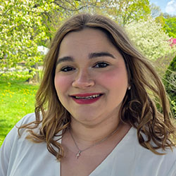 Portrait of a smiling woman with shoulder-length brown hair in a white blouse, outdoors in a sunny garden.